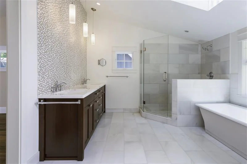 Modern bathroom with white marble tile, dark wood double vanity, two sinks, mosaic accent wall, glass-enclosed shower, and a white freestanding bathtub next to a window. Pendant lights hang above the vanity.